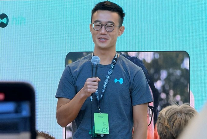 Jeff Yan, wearing a gray t-shirt with a Hyperliquid bowtie logo and a green lanyard, speaks into a microphone while discussing his vision for Hyperliquid as foundational financial infrastructure akin to AWS.