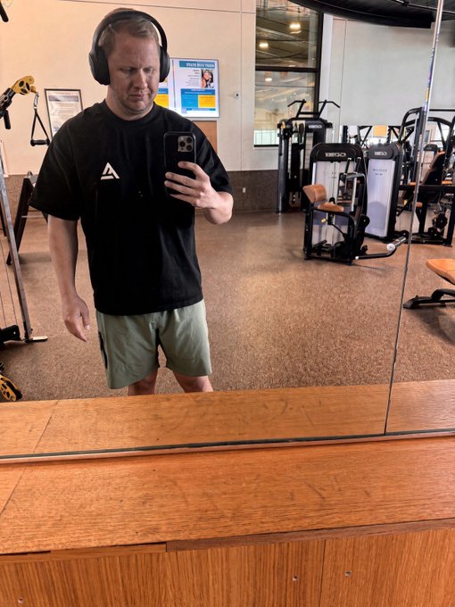 A man wearing headphones, a black t-shirt, and gray shorts takes a mirror selfie with a stern expression in a gym.
