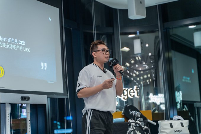 A man wearing glasses and a white t-shirt speaking into a microphone at a 'Bitget' event, with a presentation screen displaying Chinese text about '从 CEX 转账全球资产的 UEX' behind him.