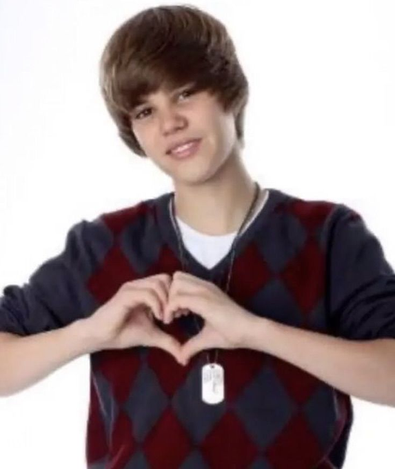 A young man with brown hair wearing a navy and maroon argyle sweater makes a heart shape with his hands against a white background.