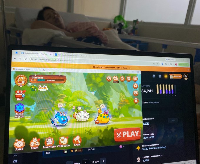 The image shows a laptop screen displaying a video game with cartoon characters, such as a cat and a dog, playing together. The person sitting next to the laptop appears to be resting or sleeping while watching the game. The scene suggests that the individual might have been spending time at home during their recovery from an illness or injury, possibly in a hospital setting.