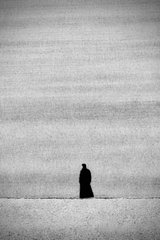A minimalist black-and-white photograph of a solitary dark figure standing on a vast, flat landscape under a hazy sky, evoking a sense of isolation.
