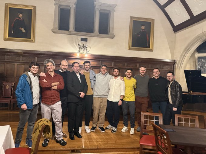 A group of eleven men poses for a group photograph in a wood-paneled hall featuring historical portraits and a grand piano.