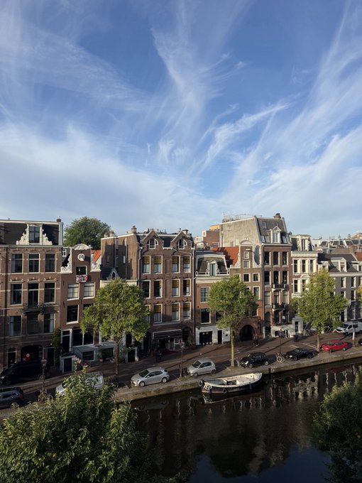 A scenic view of Amsterdam's canal-side architecture under a bright blue sky with wispy clouds, captured in the morning light. The image showcases classic Dutch townhouses with stepped gables lining the waterway, where a small boat is moored along the street, perfectly reflecting the caption's casual "GM" (good morning) greeting.