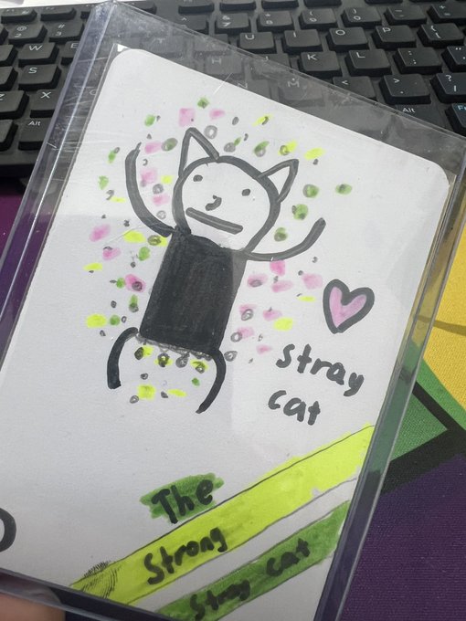 A hand-drawn artwork on a laminated card showing a smiling cat character with raised arms, surrounded by colorful dots, a pink heart, and the text 'Stray cat' and 'The Strong Stray Cat' written on it.