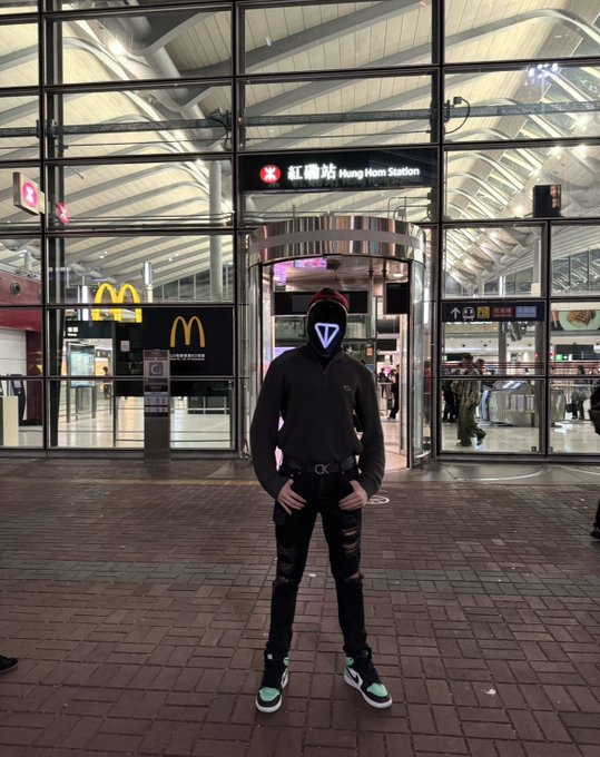A person wearing a glowing blue triangular LED mask and black hoodie stands in front of Hung Hom Station at night.