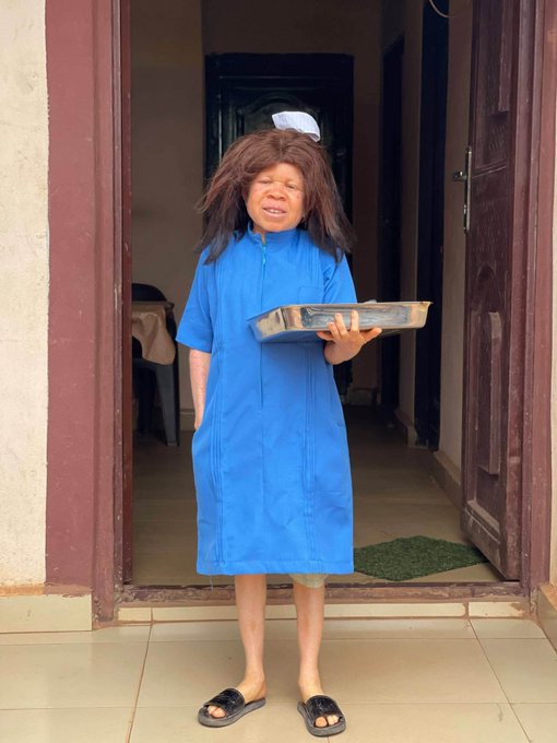 A person wearing a messy brown wig and blue robe holding a tray, standing in a doorway in a comedic pose.