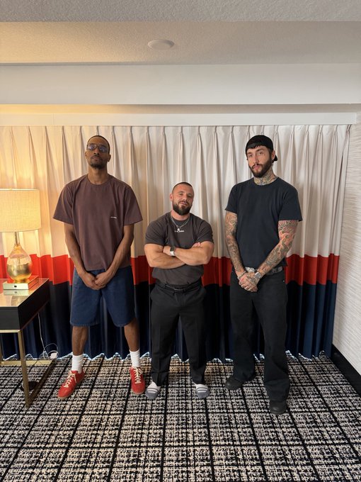 Three men pose for a photo in what appears to be a hotel room, dressed casually with one notably wearing red Jordans, capturing a relaxed group shot of friends hanging out together.