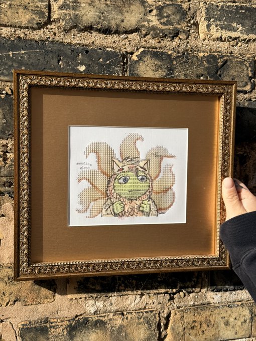A framed cross-stitch artwork of a stylized character with flame-like hair held against a brick wall, with 'Sanity's Edge' text visible.