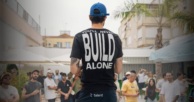 A man wearing a black t-shirt with "YOU'LL NEVER BUILD ALONE" and a "talent" logo stands with his back to the camera, facing a gathered crowd at an outdoor event, visually embodying the team spirit celebrated in the post about Talent Protocol.