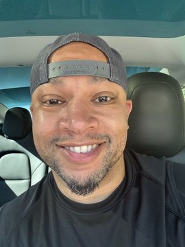 A selfie of a smiling man wearing a gray backwards cap and a black shirt, taken inside a vehicle.