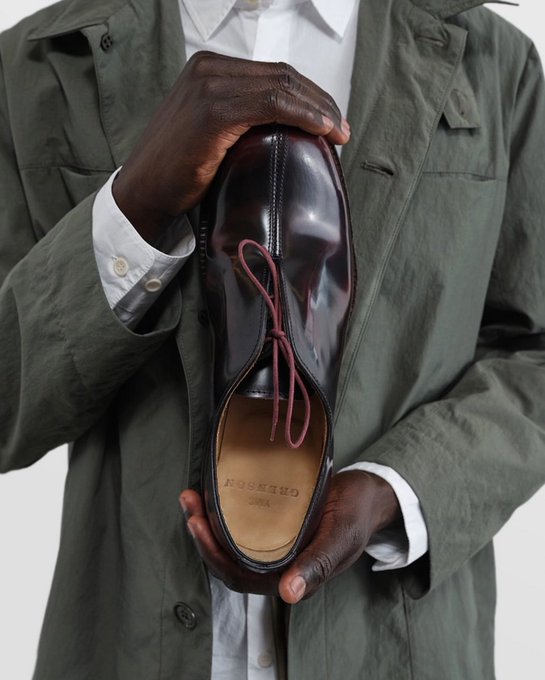 A close-up product shot features a person wearing an olive green jacket holding up a polished burgundy leather oxford shoe with red laces, branded 'GRenson' inside.