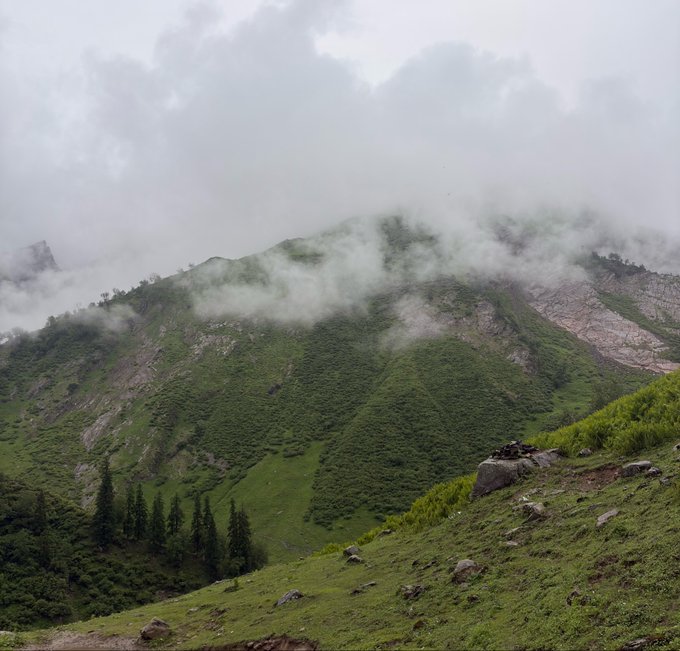 A misty, lush green mountain landscape with low-hanging clouds partially obscuring the rocky peak under an overcast sky.