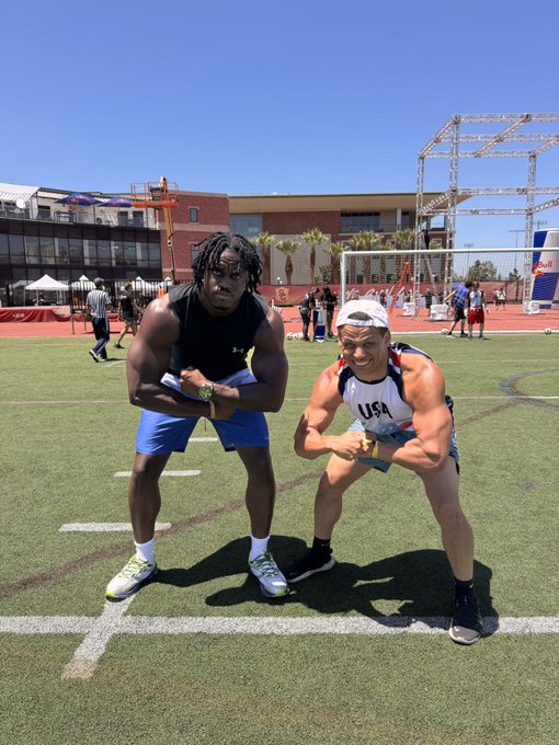 Two muscular men flex on a football field—Rams running back Todd Gurley in blue shorts and a fellow athlete in a "USA" jersey—both posing playfully under bright sun, perfectly capturing the "billions of LPs" (life points) bravado of two elite athletes joking about their combined strength.