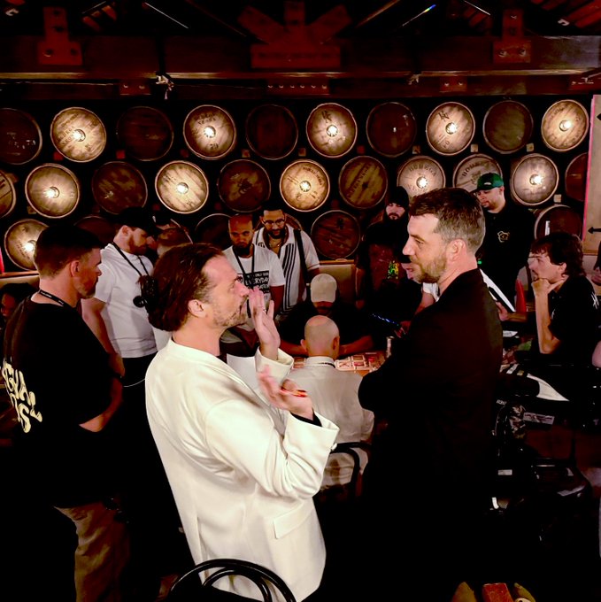 A man in a white suit talks with another man in a dark jacket inside a venue with numerous wooden barrels stacked on the wall behind them.
