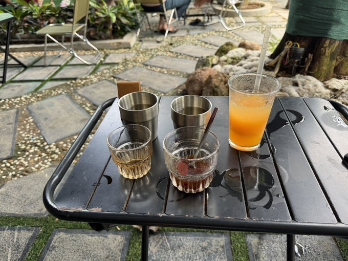 A black outdoor table holds two metal cups, two small glass cups with a dark beverage, and a plastic cup with an orange drink and straw, set in a stone-paved garden area.