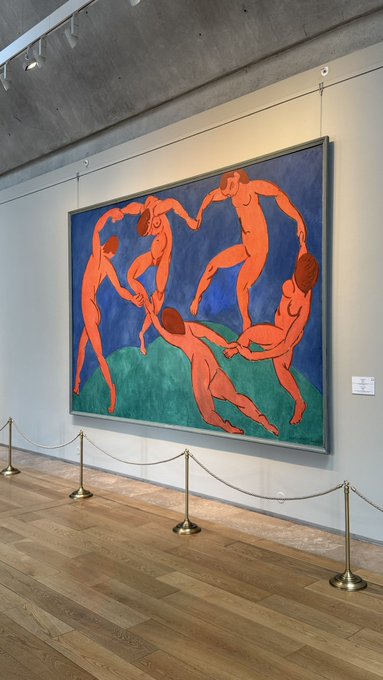 The image features a large painting of nude figures posed in various positions, with one figure holding another's hand. The painting is displayed prominently against a white wall, which contrasts with its vibrant colors. A gold chain can be seen hanging from the ceiling above the artwork. In addition to the painting itself, there are two small statues placed on either side of it, adding to the overall visual appeal and artistic presentation of the scene.