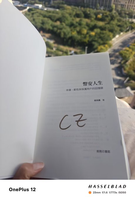 A photo taken with a OnePlus 12 (Hasselblad camera, 23mm f/1.6) showing an open book with Chinese text and a handwritten 'CZ' on the page, with an outdoor view in the background.
