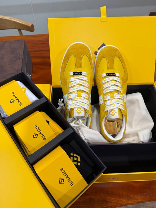 A pair of yellow sneakers with 'BINANCE' branding on the laces sits in a yellow presentation box next to matching yellow socks with the Binance logo.