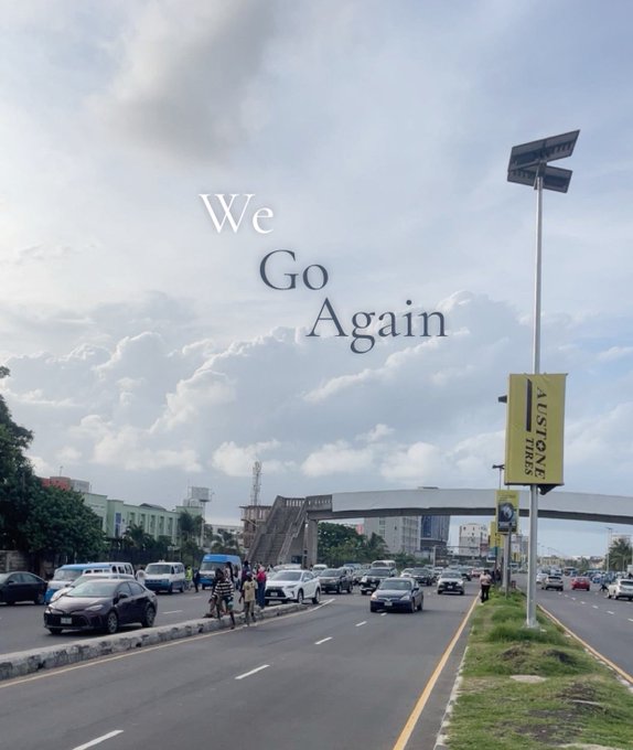 A cloudy-day street scene with traffic on a multi-lane road, featuring an overpass, vehicles, pedestrians, a yellow 'AUSTIONE TYRES' billboard, and the overlaid text 'We Go Again'.