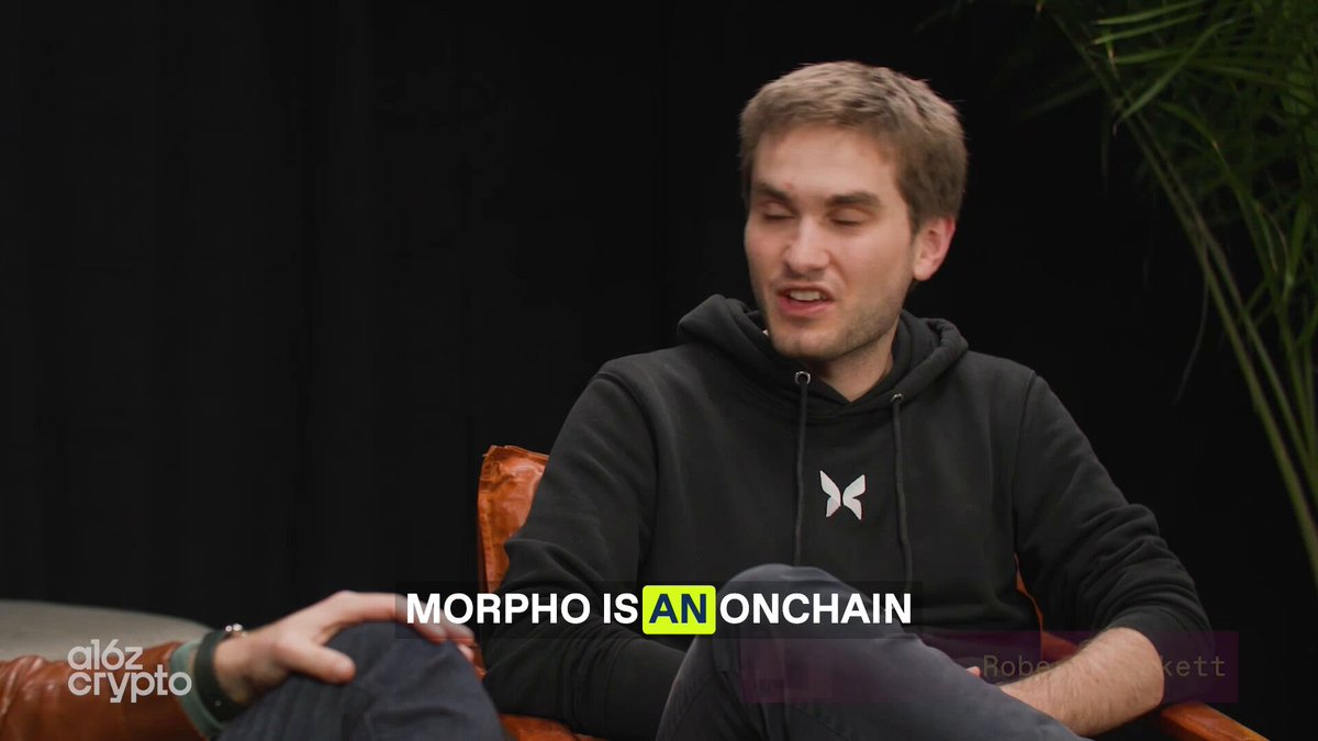 The image features a man sitting down, wearing a black hoodie with white lettering that says " Morpho" on his chest. The background of the image is dark, which contrasts with the bright yellow text written below him. Below the text, there are two lines of writing that read "What is Morpho? Cryptocurrency is about exchanging money without banks." This suggests that the man might be discussing the concept of cryptocurrency and how it works in relation to Morpho's name.