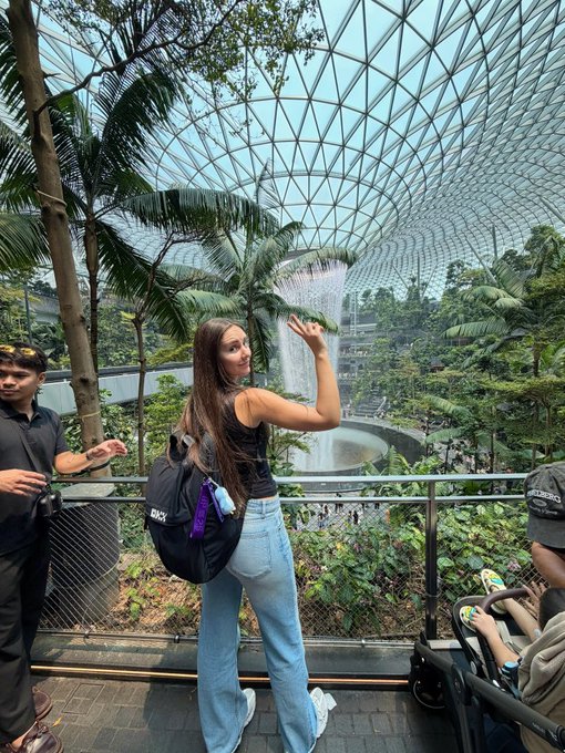 A woman poses in front of the Rain Vortex at Jewel Changi Airport, surrounded by tropical plants and a glass dome ceiling.