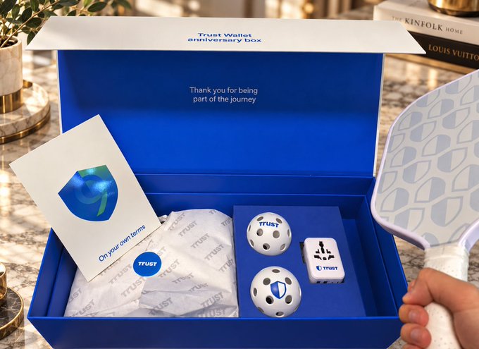 A blue "Trust Wallet anniversary box" is displayed open, featuring the text "Thank you for being part of the journey" and "On your own terms," along with branded golf balls, a Trust Wallet poker chip, a mini deck of cards, and a patterned paddle held by a person—showcasing the giveaway prize set celebrating Trust Wallet's 9th anniversary.