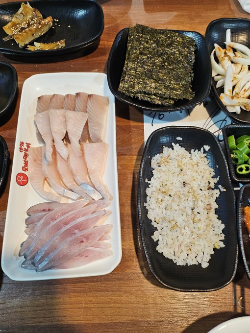 A Korean restaurant meal featuring sliced raw fish on a long plate, topped with rice and sesame, along with various banchan side dishes on the side.