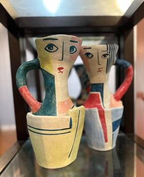 Two ceramic decorative pitchers or vases with stylized human faces, painted in cream, teal, coral, and blue tones with handle designs, are displayed on a glass shelf.