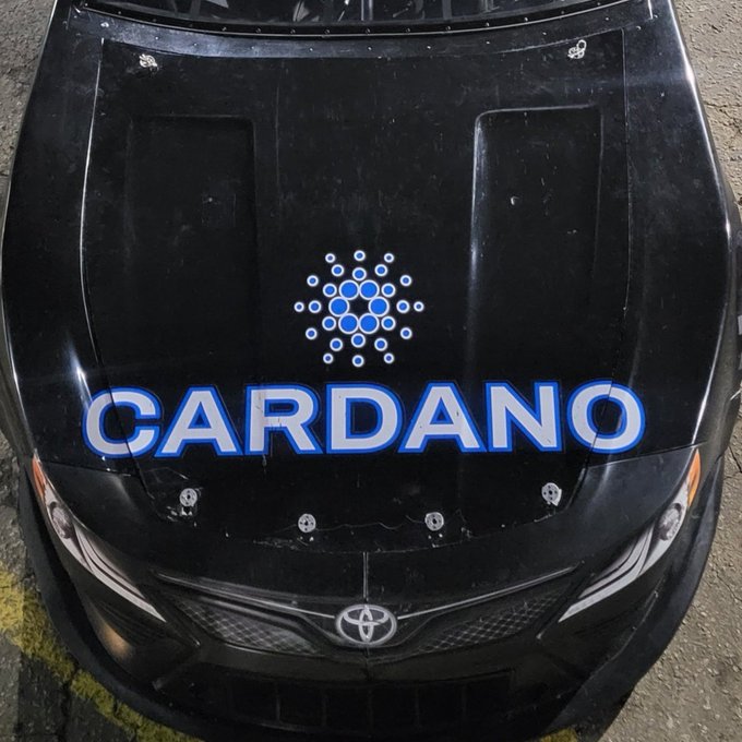 The hood of a black Toyota car featuring a 'CARDANO' decal with the blue Cardano blockchain logo.