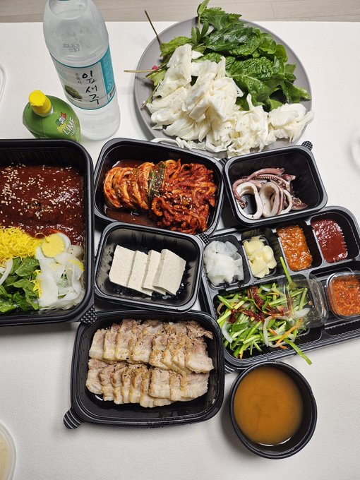 An overhead view of a Korean meal spread featuring various banchan (side dishes), boiled pork slices, tofu, vegetables, sauces, and bottles of Korean beverages.