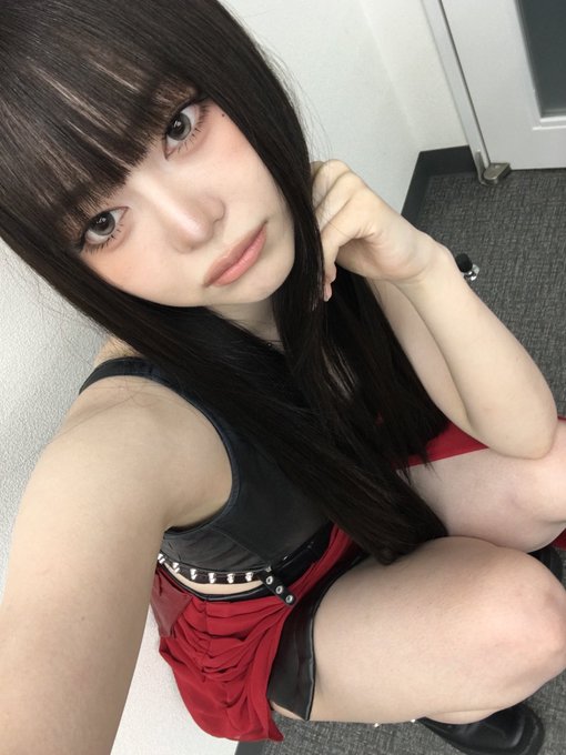 A selfie of a young woman with long dark hair and bangs wearing a black top and red skirt, crouching indoors on carpet.