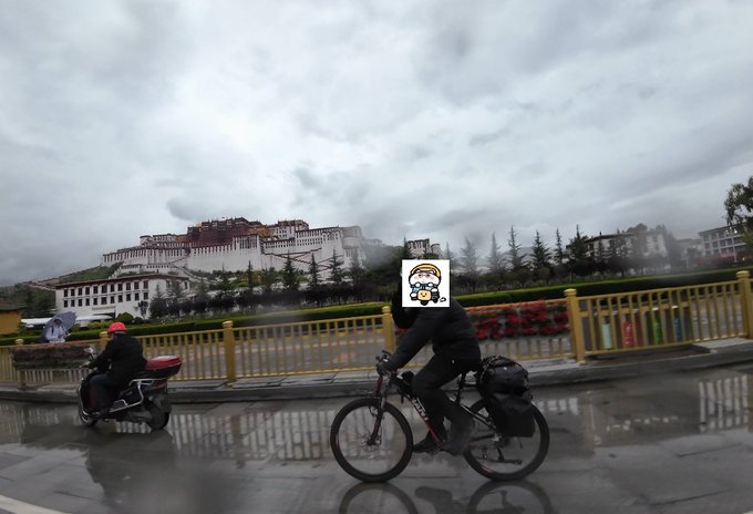 A cloudy, overcast street scene with a cyclist and a person on a red scooter riding past a yellow fence, with a large white palace-like building visible in the background mountains.