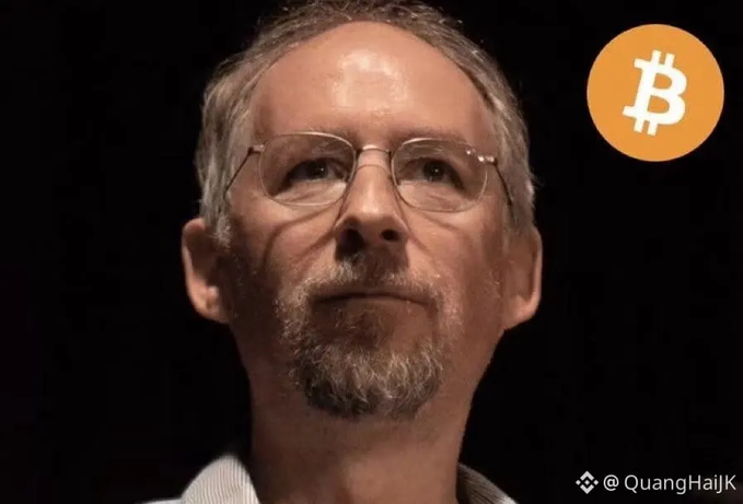 A close-up portrait of a gray-haired man with glasses and a goatee against a black background, with a Bitcoin logo in the upper right and the watermark '@QuangHaiJK'.