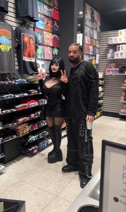 Two people pose for a photo inside what appears to be a band merchandise store, with one person wearing gothic-style clothing with horns and the other in all black.