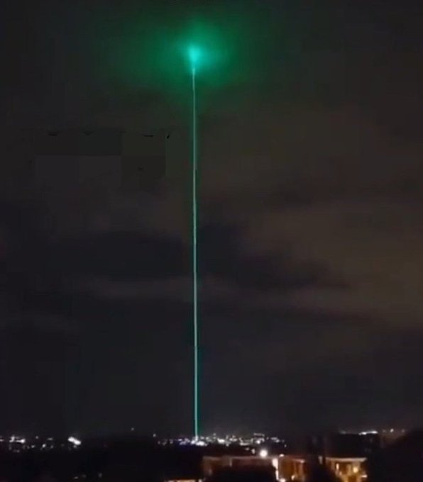 A nighttime photograph of a bright green laser beam shooting vertically into the dark sky above a cityscape.