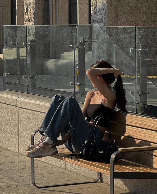 A young woman in a black top and jeans relaxes on a wooden bench, adjusting her ponytail with a black handbag beside her.