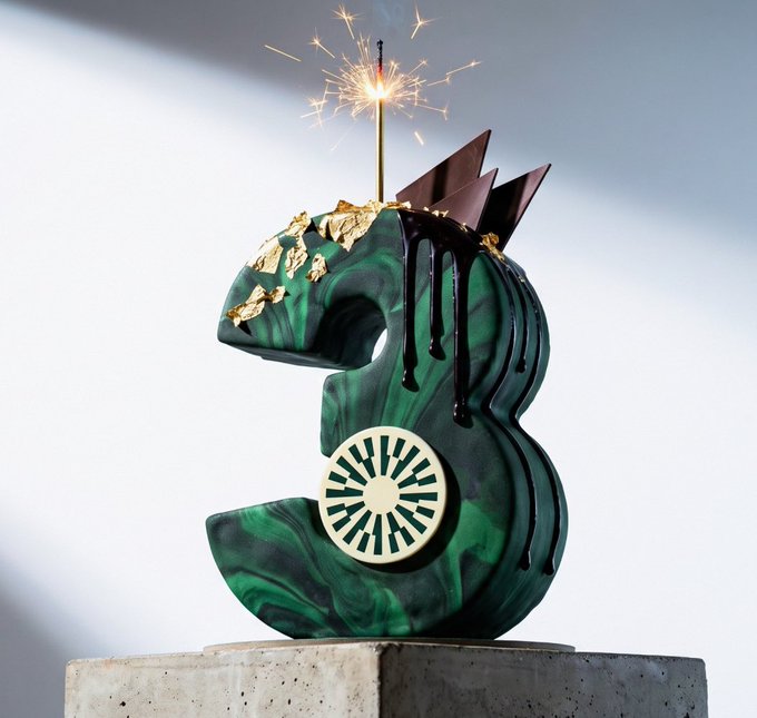 A green marbled number '3' cake with gold leaf accents, dark chocolate drips, and a lit sparkler on top, displayed on a concrete pedestal.