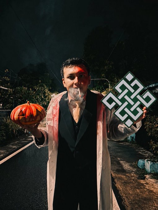 A nighttime photo of a man in a bloody lab coat smoking a cigarette, holding a carved jack-o'-lantern in one hand and a green Celtic knot prop in the other.