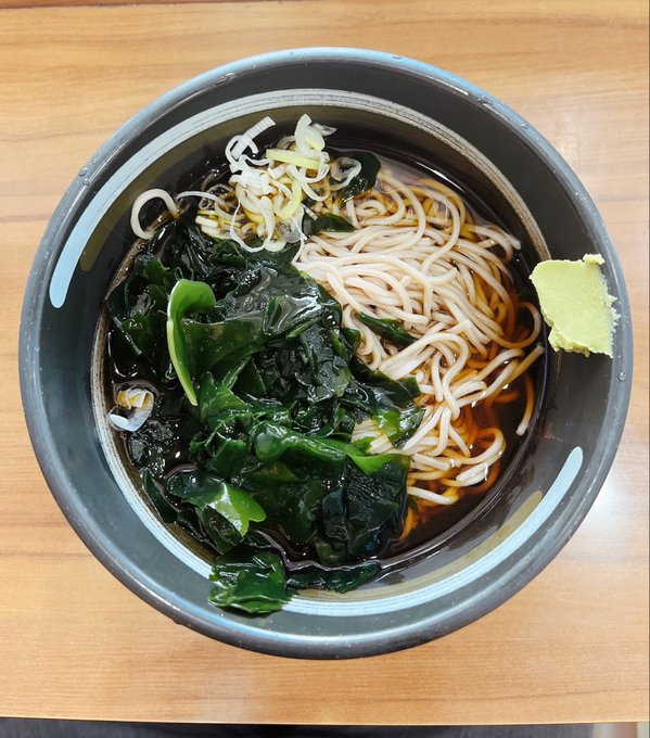 A bowl of noodle soup with dark broth, green leafy vegetables (likely seaweed or spinach), noodles, shredded toppings, and a side of green paste (possibly wasabi or ginger).