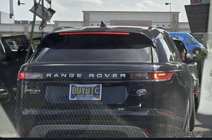 A black Range Rover Velar is shown from the rear, featuring a license plate reading "BUYBTC" with "BEDFORD" at the bottom. The custom plate cleverly echoes the post's message "BUY BTC" (Bitcoin), turning the vehicle's identification into a visible endorsement of the cryptocurrency.