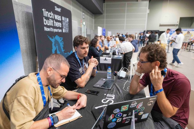 Several people wearing lanyards sit at a table with laptops during a tech event; a banner reads '1inch was built here Now it's your turn'.