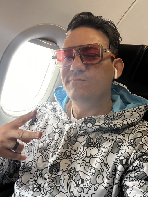 A man poses for a selfie on an airplane, wearing tinted orange sunglasses and a white hoodie with black doodle patterns, making a peace sign.
