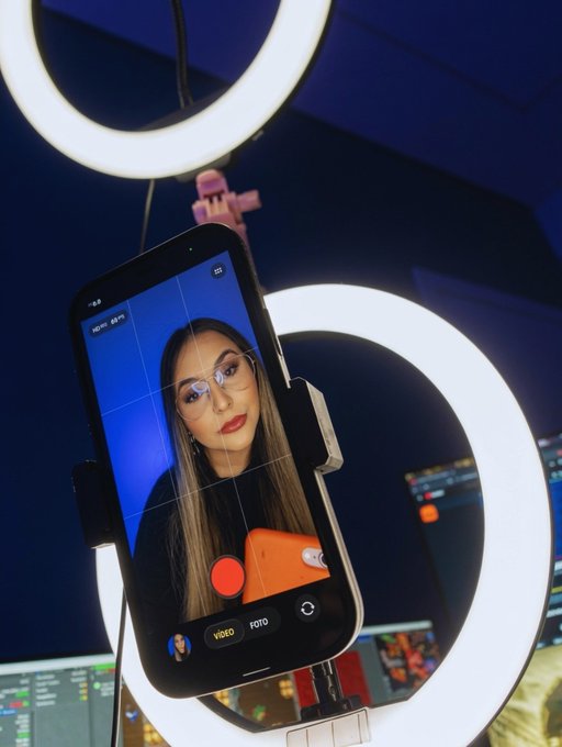 A smartphone on a tripod with a ring light displays a camera app recording a young woman wearing glasses and bold red lipstick, with "VIDEO" and "FOTO" mode options visible. The setup suggests content creation during off-peak times—mirroring the post's message that building one's reputation and craft in quiet markets pays off once the spotlight arrives.