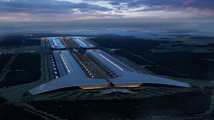 An aerial rendering of a massive futuristic airport terminal at dusk with wings-like architecture stretching across the landscape.