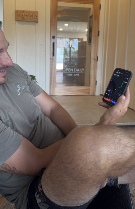A man sits casually with legs crossed while holding up his phone, which displays a trading screen showing a position in "$69Eleven" with green gains. In the background, a glass door reads "OPEN DAILY 7AM-6PM," suggesting the photo was taken at a café or shop where he's checking his first-ever meme coin investment.