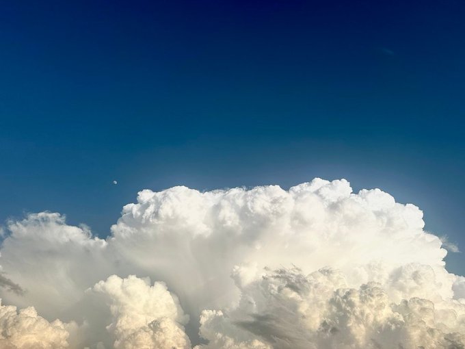 A photograph captures a dramatic, towering cumulus cloud formation against a deep blue sky, with the sun illuminating the white billowing tops.
