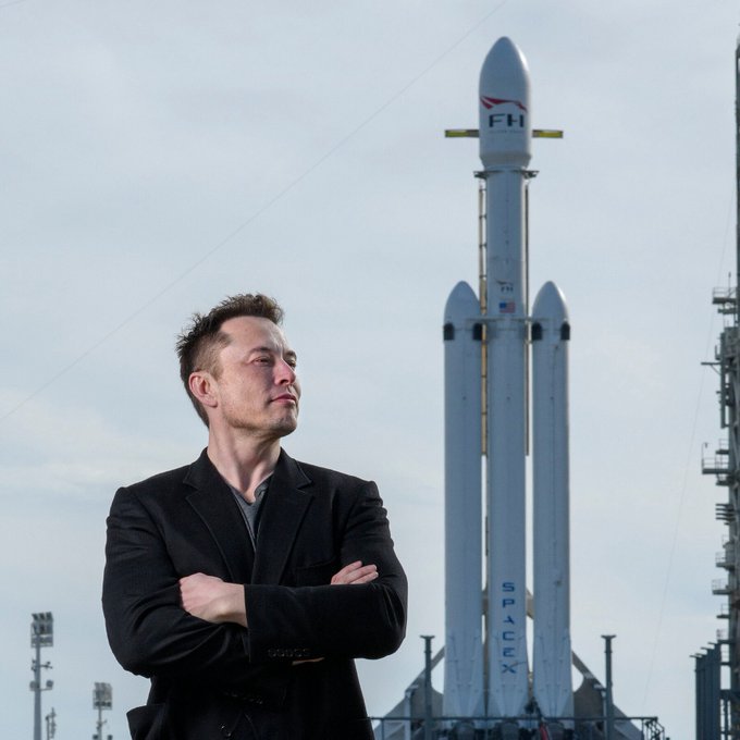 Elon Musk stands confidently with arms crossed in front of a SpaceX Falcon Heavy (FH) rocket, which prominently displays the "SPACEX" and "FH" branding along with an American flag, celebrating his 55th birthday as the visionary leader behind the aerospace company.