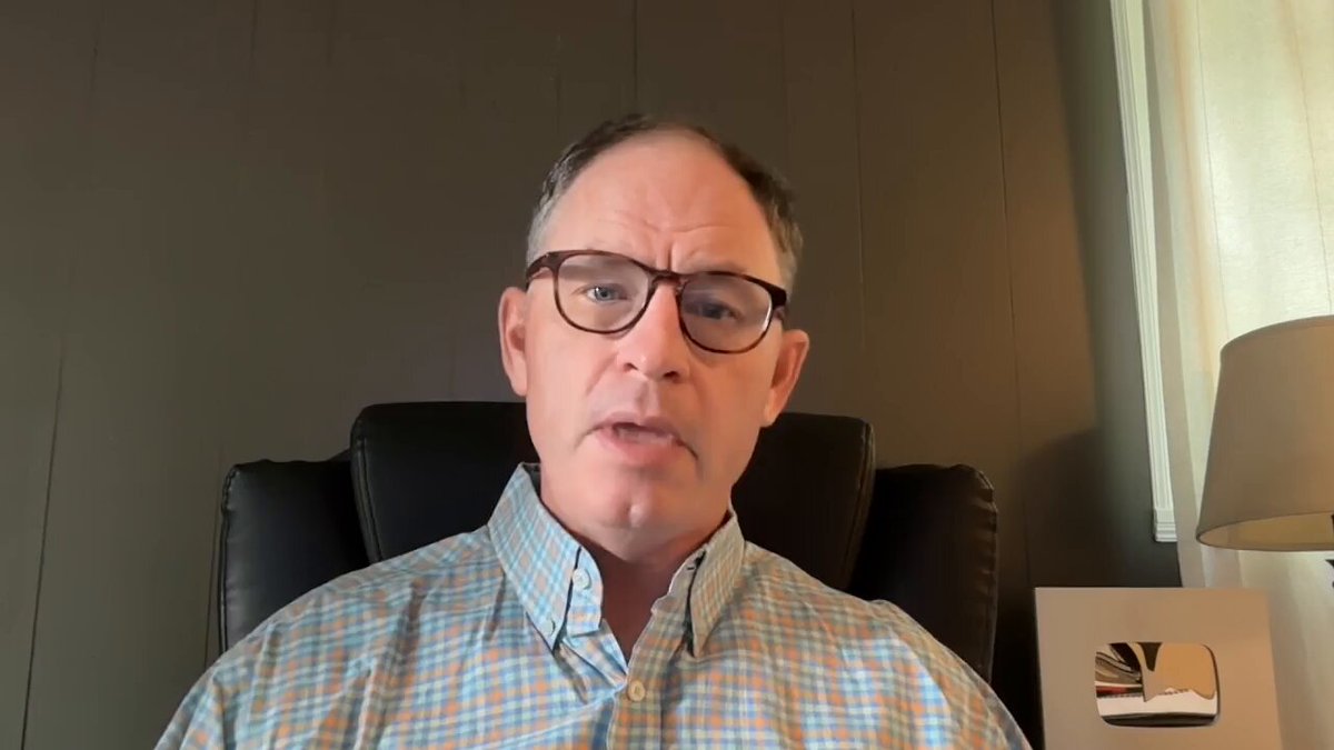The image shows a man wearing glasses sitting in an office chair, with his face clearly visible against a dark background. The man appears to be looking directly at the camera while seated. In front of him, there is a lamp that illuminates the area around him. On the right side of the image, there is a small video playing on a screen or TV. The post includes text that reads "The XRP Dip Secret Watch The Full Youtube Video Here: youtu.be/Dt9uE-KHGao". This suggests that the man in the office chair might be discussing something related to cryptocurrency or watches a video about it, possibly from a source like You Tube.