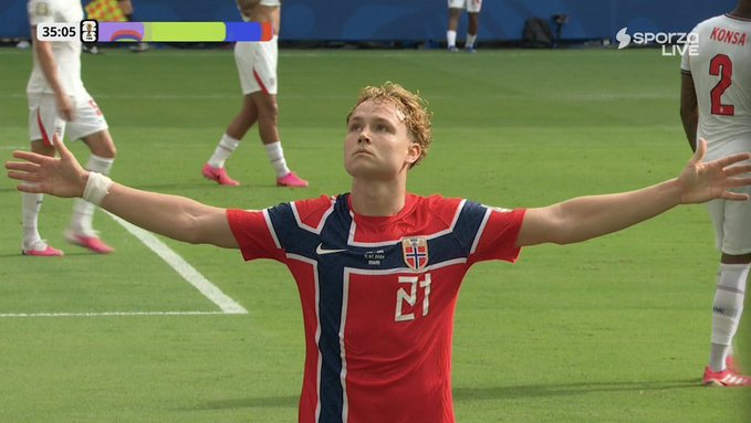 A soccer player wearing a red Norway jersey with the number 21 stands on a field with his arms outstretched and eyes closed; the scoreboard in the upper left shows '35:05' with a score of 2-2, and 'SPORZA LIVE' is visible in the upper right corner.